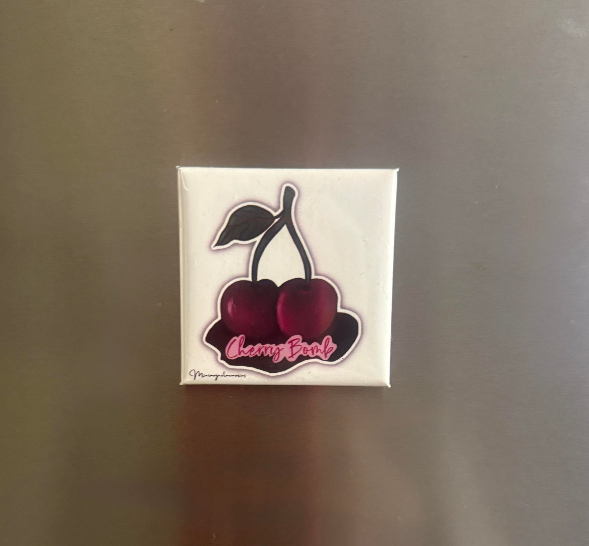 Cherry Bomb magnet with a double cherry illustration and pink script, displayed on a fridge.