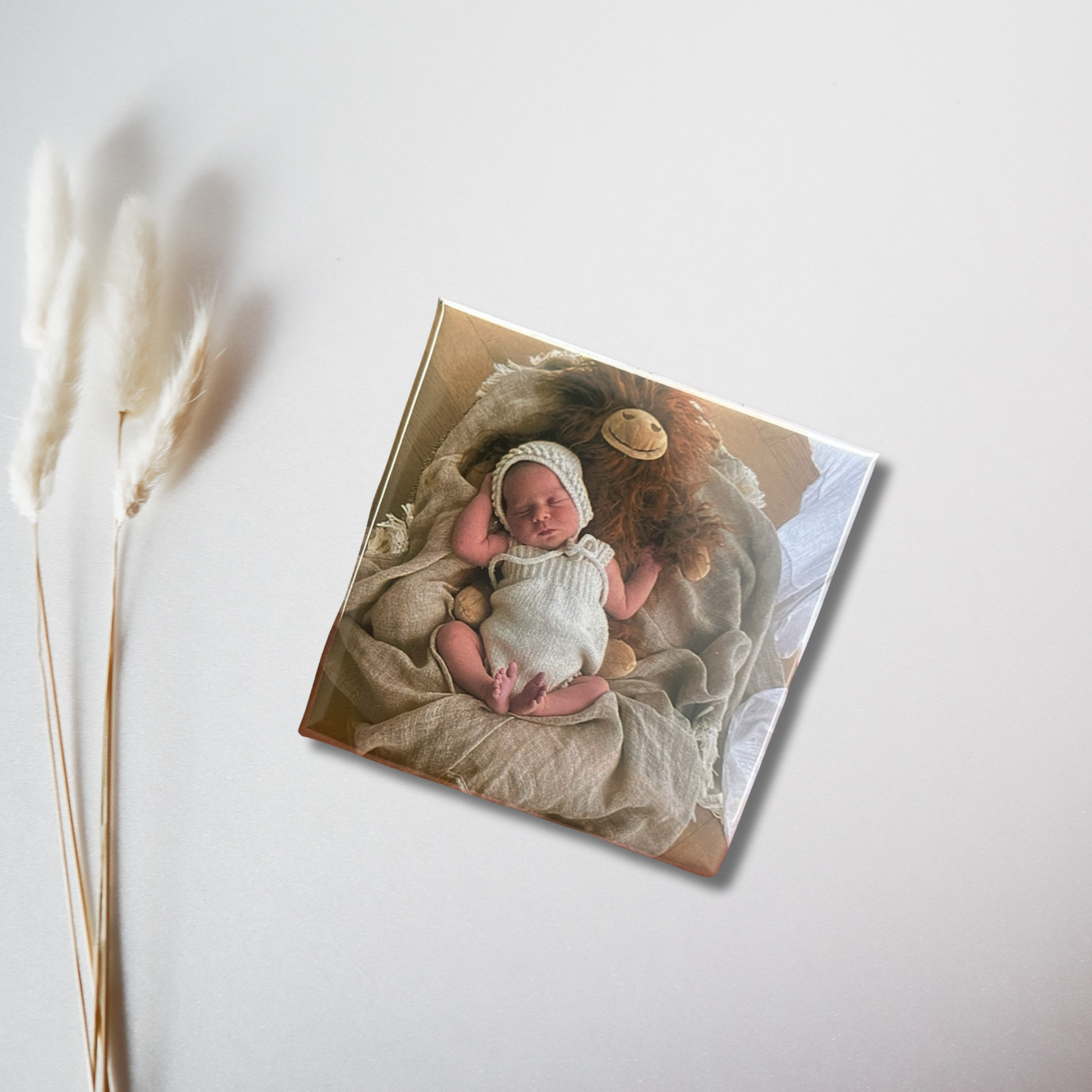  Newborn baby in a white knitted outfit sleeping on a beige blanket beside a plush lion toy, printed on a photo magnet block.